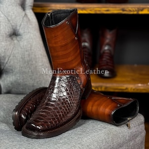 Può includere: Stivali in pelle marrone con tomaia strutturata marrone scuro e gambale liscio marrone chiaro. Gli stivali hanno una chiusura a zip e sono appoggiati su una superficie imbottita grigia. Il testo "MenExoticLeather" è visibile.