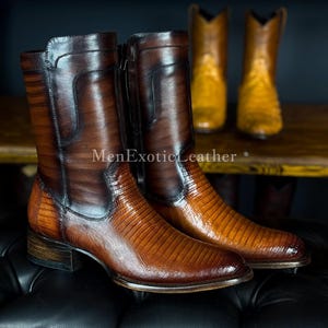 May include: A pair of brown leather boots with a textured pattern and a zipper closure. The boots are displayed on a black surface, with a pair of cowboy boots in the background. The text "MenExoticLeather" is visible.