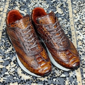 May include: Brown leather sneakers with a reptile skin design. The trainers feature brown laces, white soles, and black detailing. The interior is lined with red leather. The text "Men Exotic Leather" is visible on the side.