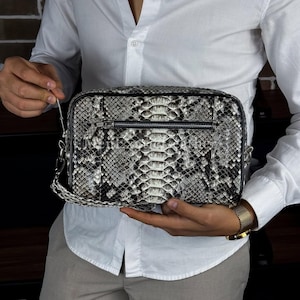 Puede incluir: Un bolso de mano rectangular con un estampado de piel de serpiente en tonos grises, negros y blancos. El bolso tiene cierre de cremallera y un pequeño bolsillo frontal. Incluye una correa de hombro desmontable. El bolso lo sostiene una persona con una camisa blanca.
