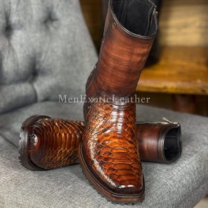 Può includere: Un paio di stivali in pelle marrone con un motivo strutturato e squamoso nella parte inferiore e pelle liscia sullo stelo superiore. Gli stivali hanno cerniere e sono appoggiati su una superficie imbottita grigia. Il testo "MenExoticLeather" è visibile.