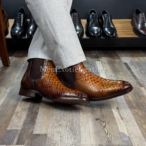May include: Brown leather Chelsea boots with a textured pattern. The boots have a pointed toe and a low heel. Other dress shoes are visible in the background. The text "MenExoticLeather" is visible at the bottom of the image.