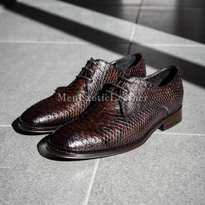May include: A pair of brown leather dress shoes with a snakeskin pattern. The shoes are laced up and have a low heel. The text "Men Exotic Leather" is visible on the side of the shoes.