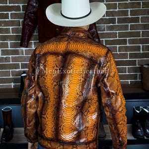 Op de afbeelding: Een bruin en oranje leren jasje met slangenpatroon en een witte cowboyhoed. De jas heeft een ritssluiting en een geribbelde zoom. De tekst "MenExoticLeather" is zichtbaar op de jas.