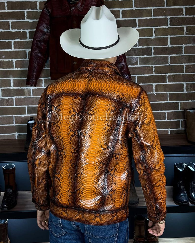 Op de afbeelding: Een bruin en oranje leren jasje met slangenpatroon en een witte cowboyhoed. De jas heeft een ritssluiting en een geribbelde zoom. De tekst "MenExoticLeather" is zichtbaar op de jas.
