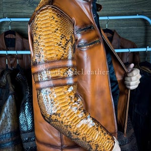 May include: A brown leather jacket with snake skin patterned sleeves and shoulder accents. The jacket has a stand-up collar, two zippered pockets, and a front zipper closure. The sleeves have three horizontal bands. The jacket is displayed on a rack with other jackets.