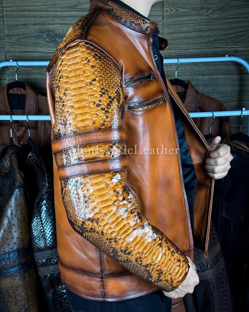 May include: A brown leather jacket with snake skin patterned sleeves and shoulder accents. The jacket has a stand-up collar, two zippered pockets, and a front zipper closure. The sleeves have three horizontal bands. The jacket is displayed on a rack with other jackets.