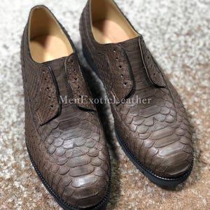 May include: A pair of brown leather dress shoes with a textured, scale-like pattern. The shoes have a classic design with a rounded toe and a low heel. The text "MenExoticLeather" is visible on the shoes.