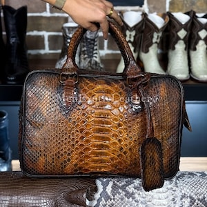 May include: A brown and tan leather travel bag with a reptile skin pattern. The bag has a top handle, a luggage tag, and the text "Men Exotic Leather" is visible. The bag is held by a person.