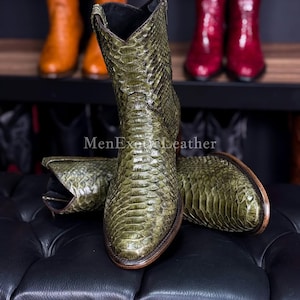 Puede incluir: Botas de vaquero verde oliva con un patrón de piel de serpiente, expuestas sobre una superficie de cuero negro. Las botas tienen una punta puntiaguda y un tacón apilado. El texto "MenExoticLeather" es visible. Otras botas naranjas y rojas están en un estante en el fondo.
