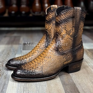 Op de afbeelding: Een paar bruin-zwarte leren cowboylaarzen met een slangenpatroon. De laarzen hebben een puntige neus, een kleine hak en een ritssluiting aan de zijkant. De laarzen worden gepresenteerd op een houten oppervlak.