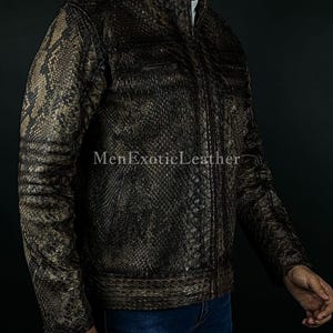 May include: A brown and tan snakeskin-patterned jacket with a zip-up front and a stand-up collar. The jacket has two chest pockets and the text "MenExoticLeather" is visible. The jacket is worn with blue jeans.