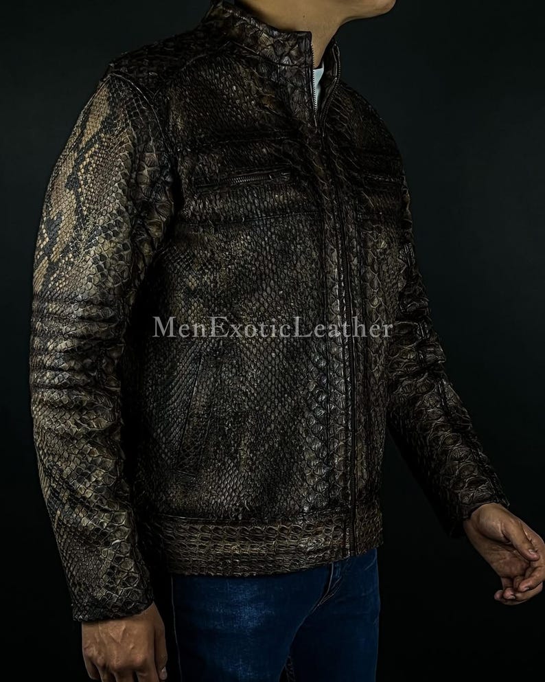 May include: A brown and tan snakeskin-patterned jacket with a zip-up front and a stand-up collar. The jacket has two chest pockets and the text "MenExoticLeather" is visible. The jacket is worn with blue jeans.