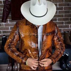 Op de afbeelding: Een bruine jas met slangenpatroon en een witte cowboyhoed. De jas heeft een knoopsluiting en lange mouwen. De tekst "MenExoticLeather" is zichtbaar op de jas. Laarzen zijn zichtbaar op de achtergrond.