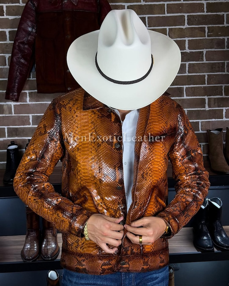 Op de afbeelding: Een bruine jas met slangenpatroon en een witte cowboyhoed. De jas heeft een knoopsluiting en lange mouwen. De tekst "MenExoticLeather" is zichtbaar op de jas. Laarzen zijn zichtbaar op de achtergrond.