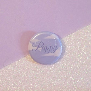 Könnte beinhalten: Ein lila und weißer Knopf mit dem Wort "Puppy" in kursiv. Der Knopf hat ein Wolken-Design.