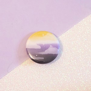 May include: A round button with a yellow, white, purple, and black design. The design resembles a sunset with clouds and stars.