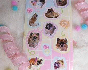 May include: A sheet of stickers featuring various images of small dogs in different poses. The stickers are on a pink and blue background with a grid pattern. The stickers are surrounded by pink and blue hearts and stars.