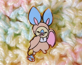 May include: A gold-colored enamel pin featuring a cartoon bat with pink, blue, and beige accents. The bat has large, rounded ears and a smiling face.