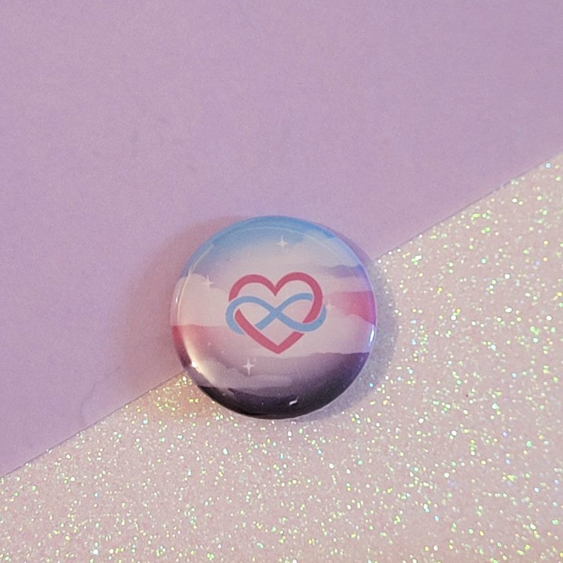 May include: A round button with a watercolor-style background in shades of blue, pink, and purple. A white infinity symbol is inside a pink heart, and the symbol is blue. The button is on a sparkly background.