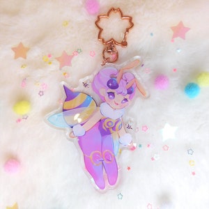 May include: A clear acrylic keychain featuring a cartoon character with purple hair, a bee costume, and a bee wings. The character is holding a beehive. The keychain is attached to a gold metal chain with a flower-shaped charm.