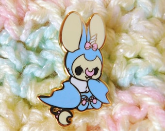 May include: A gold-colored enamel pin featuring a cartoon bat wearing a blue and white costume with a pink bow. The bat has a light yellow face with black eyes and a pink nose.