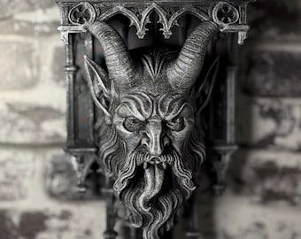 May include: A dark gray wall-mounted shelf featuring a sculpted horned creature head with a long beard and protruding tongue. The shelf has a flat top surface and is decorated with intricate gothic-style architectural arches and vertical pillars.