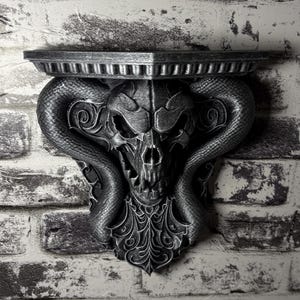 May include: A silver-colored wall shelf with a gothic design. The shelf features a skull with snake-like horns and ornate detailing. The shelf is mounted on a brick wall.