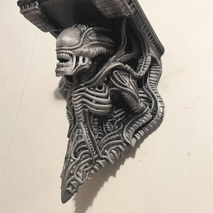 Puede incluir: Soporte de estante plateado y negro con temática Alien. El soporte presenta una escultura detallada de una criatura Alien, con un acabado metálico texturizado. El estante está diseñado para montarse en la pared, ofreciendo un elemento decorativo y funcional único.