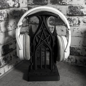 May include: A pair of white over-ear headphones resting on a black stand shaped like a gothic archway with a central pentagram design. The headphones feature a padded headband and soft ear cushions, while the stand has a rectangular base.