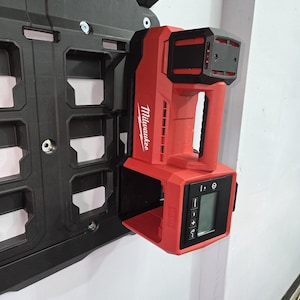 May include: A red and black Milwaukee laser level mounted on a black wall organizer. The laser level has a digital display and control buttons. The Milwaukee logo is visible on the side. The organizer has multiple square compartments.