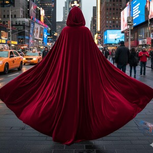 May include: A long, flowing, hooded cape in a deep red hue. The cape is made of a soft, velvety material and is spread out, creating a dramatic effect. The background shows a city street with yellow taxis and pedestrians.