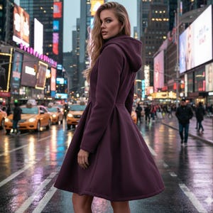 May include: A woman wearing a dark purple hooded coat with a flared skirt, standing on a wet city street. The coat has a fitted bodice and a flowing skirt. The background features city buildings, billboards, and yellow taxis.