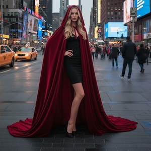 May include: A woman in a black dress and black heels stands in a city street, wearing a long, hooded, burgundy cape. The cape trails on the ground. Yellow taxis and city buildings are in the background.