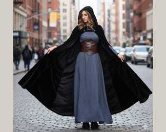 May include: A woman wearing a black hooded cloak over a long blue dress and a brown leather corset. The cloak is open, revealing the dress and corset. The background is a city street with buildings and cars.