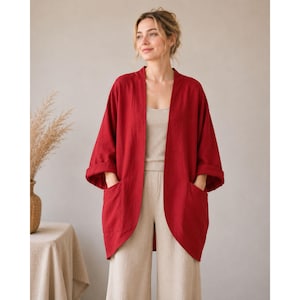 May include: A vibrant red open-front cardigan featuring three-quarter length sleeves with folded cuffs and two large patch pockets on the front. The garment has a relaxed, straight-cut silhouette and is worn over a beige tank top and matching trousers.