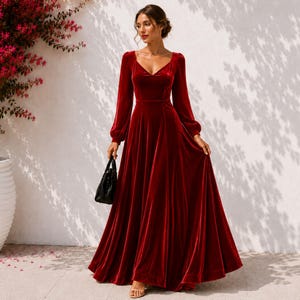 May include: A floor-length deep red velvet dress featuring long, full sleeves, a fitted bodice with a V-neckline, and a flowing, flared skirt. The garment is paired with a small black handbag held at the side, showcasing a smooth, rich texture.