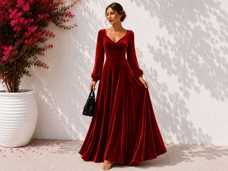 May include: A floor-length deep red velvet dress featuring long, full sleeves, a fitted bodice with a V-neckline, and a flowing, flared skirt. The garment is paired with a small black handbag held at the side, showcasing a smooth, rich texture.