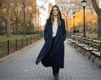 May include: A woman wearing a long, navy blue hooded coat, walking on a brick path in a park. The coat is open, revealing a cream-colored turtleneck sweater. The background features trees with fall foliage and park benches.