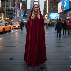 May include: A long, hooded cape in a deep red color. The cape is made of a flowing fabric and is worn by a person with long blonde hair. The background shows a city street with yellow taxis and tall buildings.