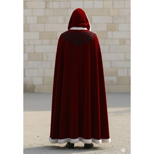 May include: A long, burgundy velvet cloak with a hood, trimmed with white faux fur. The cloak features a black embroidered design on the shoulders and a white lining in the hood. The person is standing in front of a light-colored brick wall.