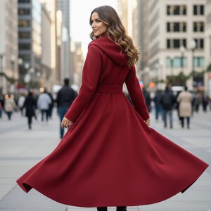 May include: A long, flowing, burgundy-colored coat with a hood and a belted waist. The coat is made of a soft material and is being worn outdoors in a city setting. The coat's skirt flares out, creating a dramatic effect.
