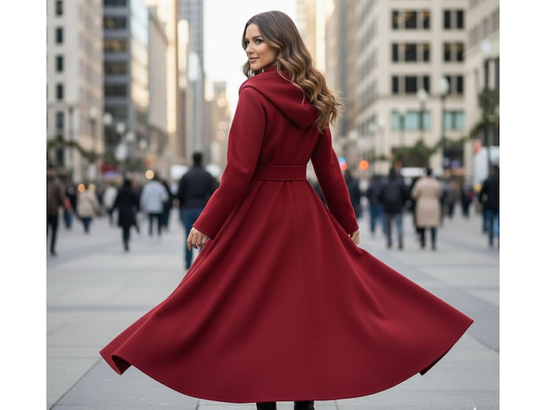 May include: A long, flowing, burgundy-colored coat with a hood and a belted waist. The coat is made of a soft material and is being worn outdoors in a city setting. The coat's skirt flares out, creating a dramatic effect.