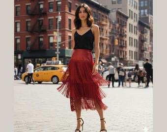 May include: A woman in a black camisole top and a red fringed skirt stands on a city street. The skirt has multiple layers of fringe. She wears black high-heeled sandals. The background includes buildings and a yellow taxi cab.