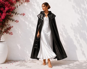Op de afbeelding: Een zwarte cape met capuchon tot op de grond, voorzien van een gestructureerde kraag en sluiting aan de voorzijde, gedragen over een mouwloze witte satijnen slipdress met een boothals. Het geheel is gecombineerd met een kleine zwarte handtas en sandalen.
