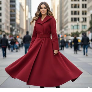 May include: A long, flowing, burgundy coat with a matching belt and hood. The coat is worn by a person in a city setting, with the coat's skirt flared out. The coat's design is elegant and classic.