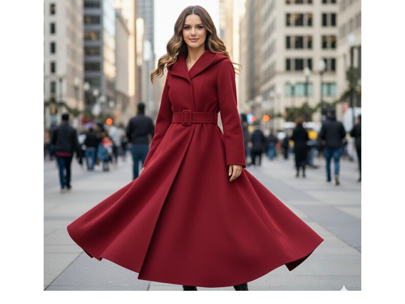 May include: A long, flowing, burgundy coat with a matching belt and hood. The coat is worn by a person in a city setting, with the coat's skirt flared out. The coat's design is elegant and classic.