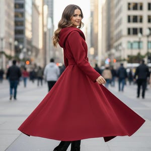 May include: A long, flowing red coat with a hood and belt, worn outdoors. The coat is a deep shade of red and has a flared skirt. The person is walking on a city street.
