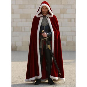 May include: A burgundy hooded cloak with white faux fur trim. The cloak is worn over a dark outfit and paired with a sword. The hooded cloak is a medieval-inspired garment.