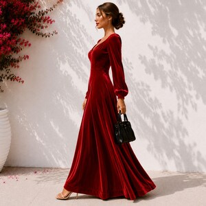 May include: A floor-length deep red velvet dress with long sleeves, a fitted bodice, and a flowing skirt. The dress features a V-neckline and cuffed sleeves. A small black handbag is held at the side, and beige strappy high-heeled sandals are worn.